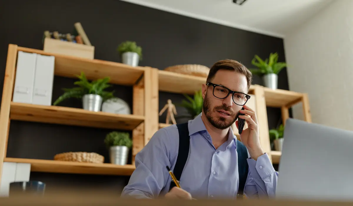 A glasses-wearing man sits talking on the phone in his home office. Article on key tools for teleworking from Bigle Legal.